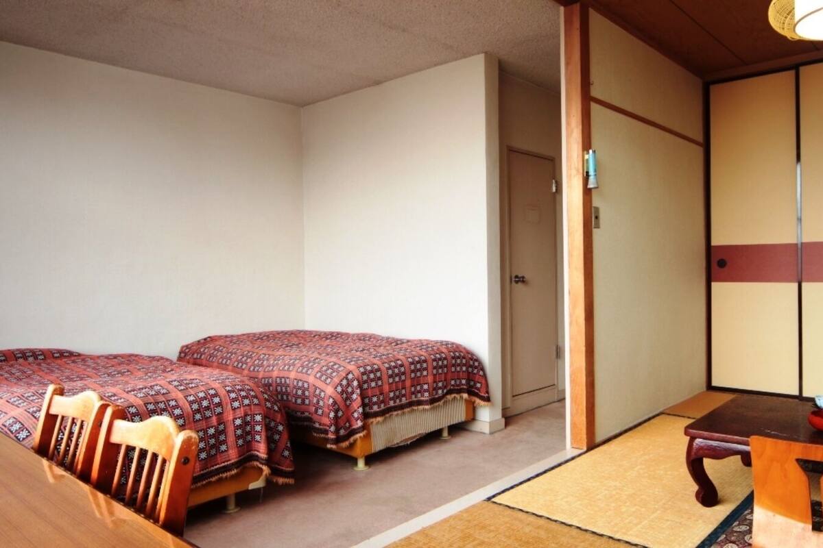 Une pièce de style japonais traditionnel, avec un sol recouvert de tatamis, deux lits avec une literie à motifs, une table en bois sur laquelle se trouve un bol, et des portes coulissantes.