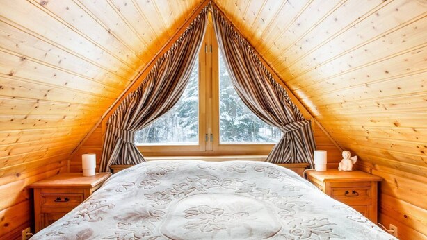 Un dormitorio acogedor en el ático, con techo inclinado, un ventanal con cortinas, una cama con ropa de cama floral y mesitas de noche de madera.