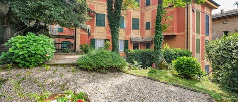 A courtyard with a gravel path, a potted plant with flowers, and a building with green shutters and a red facade.