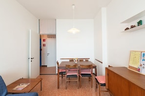 Une pièce comprenant une table à manger, des chaises, un petit meuble, un luminaire suspendu et une porte donnant sur une autre pièce.