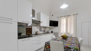 A modern kitchen with white cabinets, a built-in oven, and a dining area with a table covered in a fruit-patterned tablecloth. - Dimora del Delfino Curioso (Lecce)