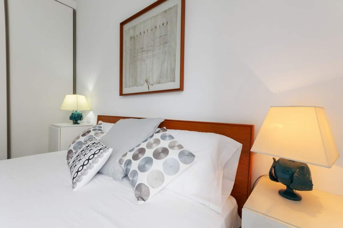 A neatly made bed with patterned pillows, a framed document on the wall, and two bedside lamps.