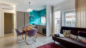 A modern living room with a sofa, a dining table with chairs, a kitchen area with a window, and a red patterned rug. - Inner City Apartments (Lecce)
