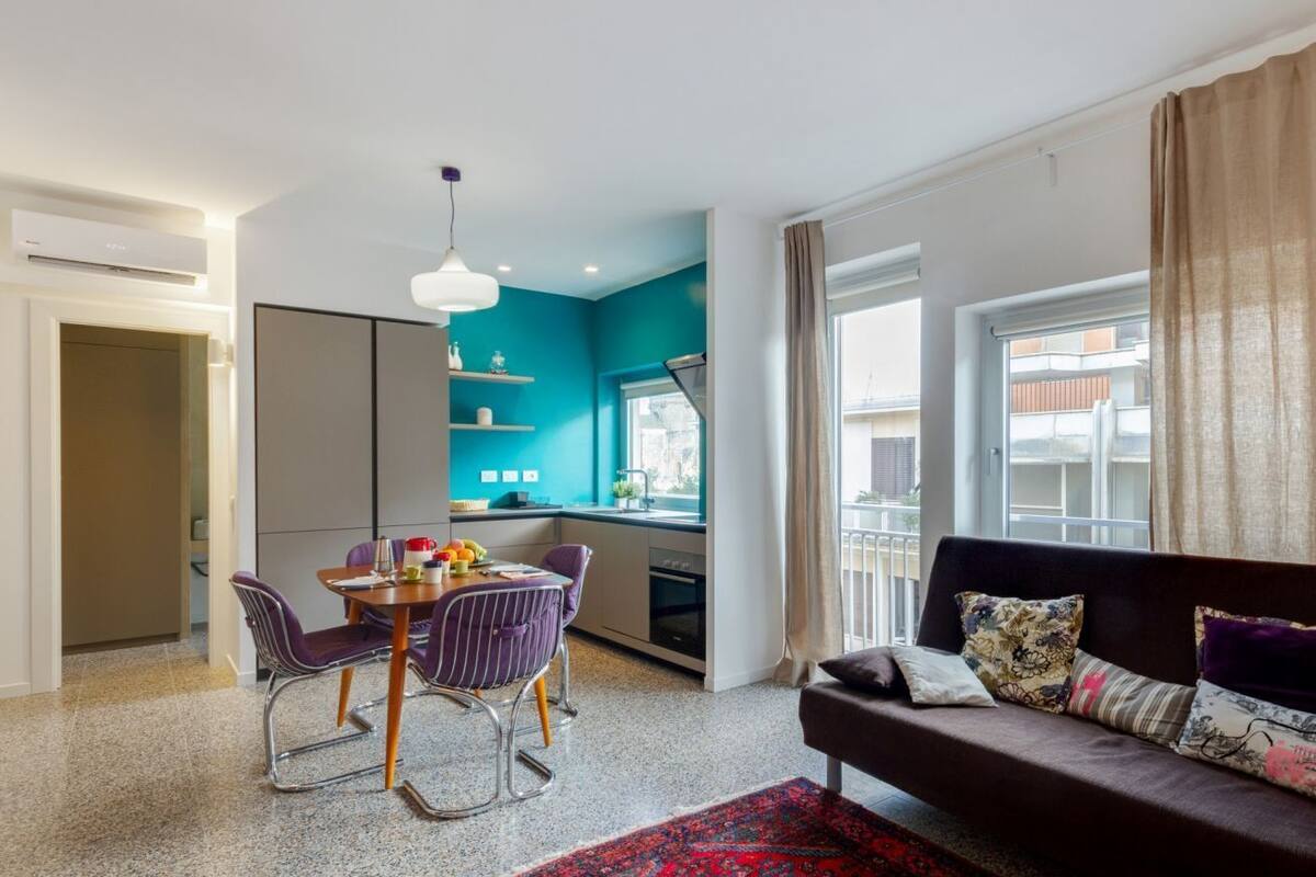 A modern living room with a sofa, a dining table with chairs, a kitchen area with a window, and a red patterned rug.