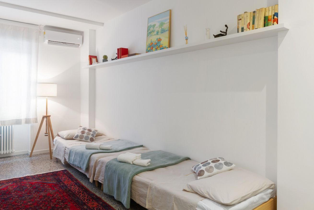 A minimalist bedroom with two single beds, a red patterned rug, a standing lamp, and a shelf with books and decorative items.