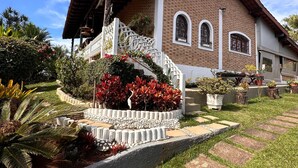 A two-story house with a brick facade, a red roof, and a white porch. There is a small garden with red flowers and a white fence. - Country house in Atibaia for rent with swimming pool with solar heating and Wi-Fi. (Atibaia)