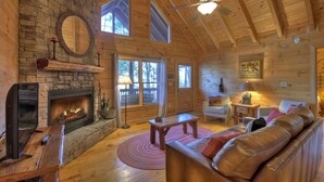 A log cabin interior with a stone fireplace, wooden furniture, and large windows. - Lakeside Cabin w/ mountain views, dock, fire pit, hot tub, and much more! (Ellijay)
