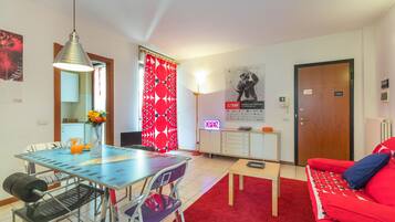 A furnished living room with a red sofa, a dining table with chairs, a coffee table, a red patterned curtain, and a poster on the wall.