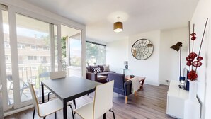 Een moderne woonkamer met een eethoek, grote ramen, een ronde klok aan de muur en een decoratieve rode plant.