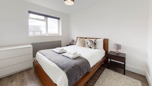 Een slaapkamer met een houten bed, witte beddengoed, een grijze plaid, een bedlampje en een raam met zonwering.