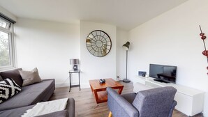 Een moderne woonkamer met een grijze bank, een grijze fauteuil, een houten salontafel, een televisie op een witte standaard en een vloerlamp.