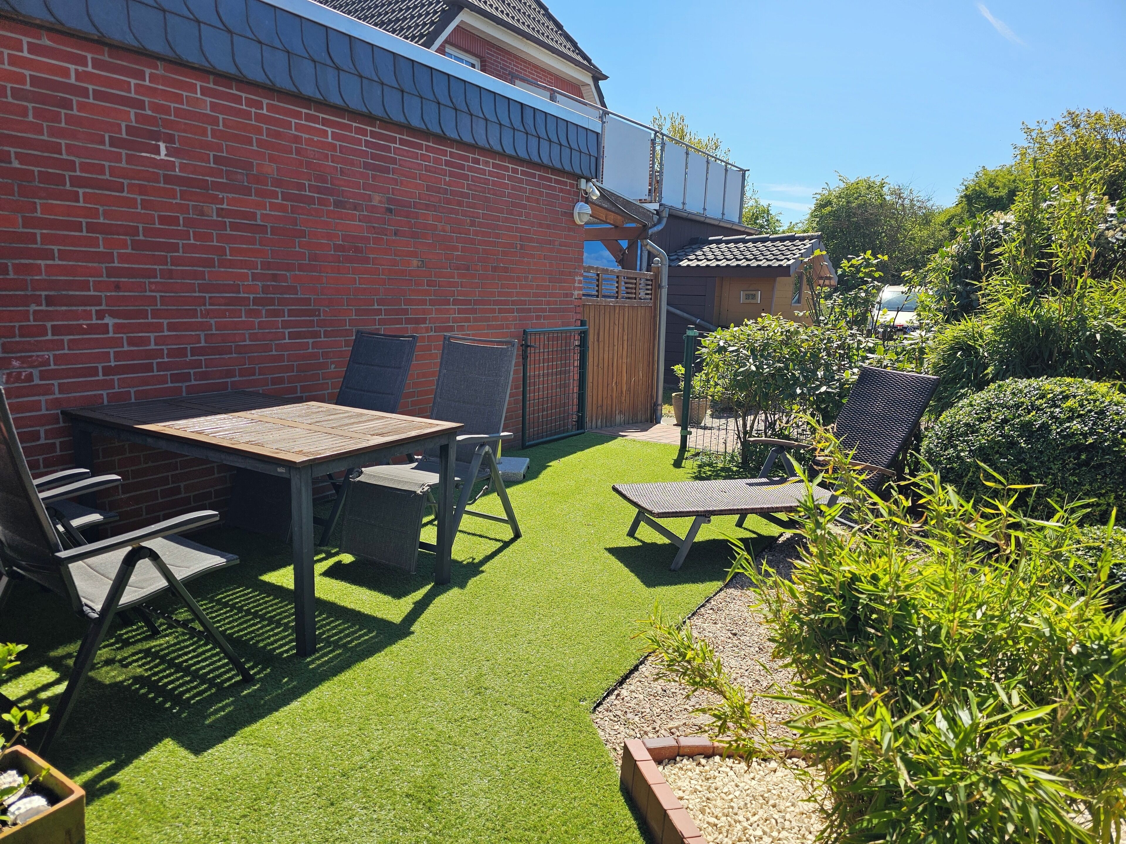 Une terrasse avec une table et des chaises, un mur en briques et un petit jardin agrémenté de plantes et d’une clôture en bois.