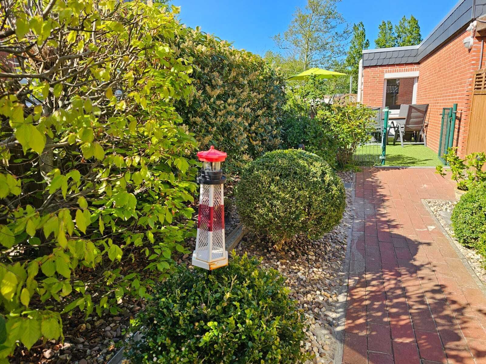 Ein Gartenweg mit einer Laterne, dichtem Grünwerk und einem Backsteinhaus im Hintergrund.