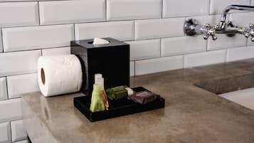 A bathroom with a white tiled wall, a black tissue box, a roll of toilet paper, and a small tray with toiletries.