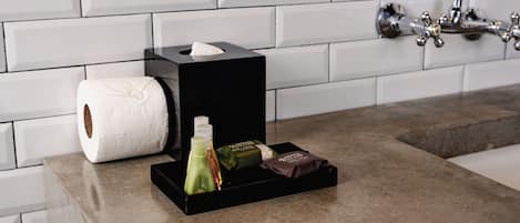 Un baño con pared de azulejos blancos, una caja de pañuelos negros, un rollo de papel higiénico y una pequeña bandeja con artículos de aseo.