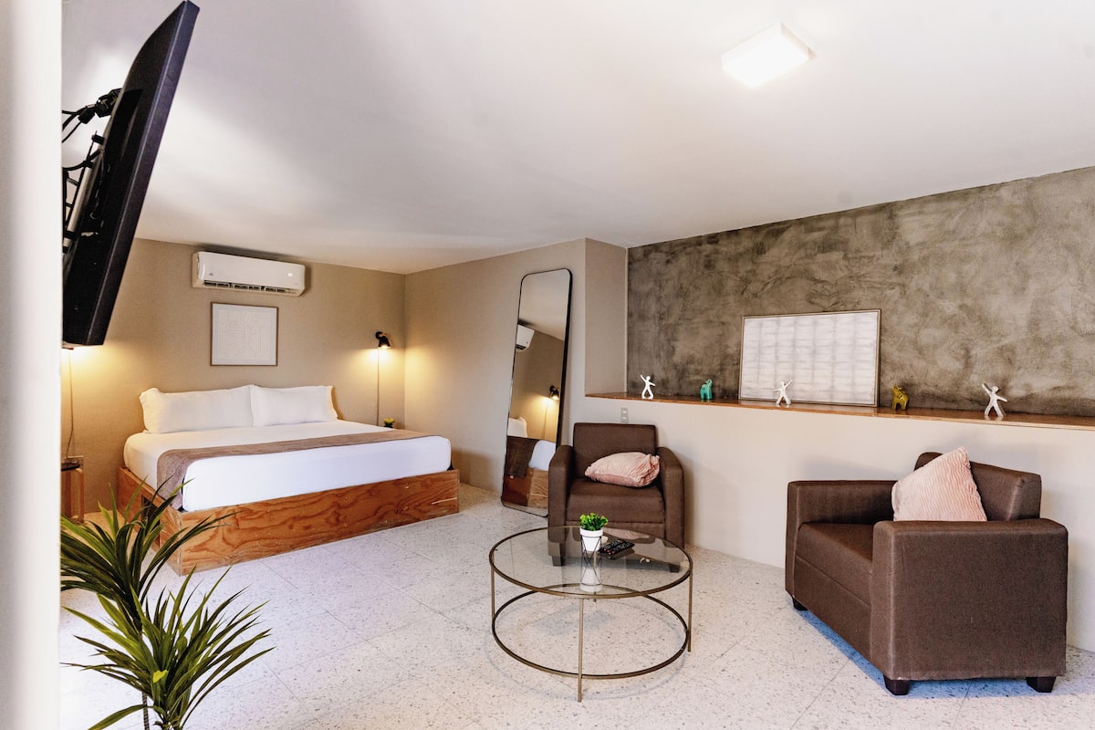 A hotel room with a bed, a sofa, a chair, a round coffee table, a TV, a mirror, and a wall-mounted air conditioner.