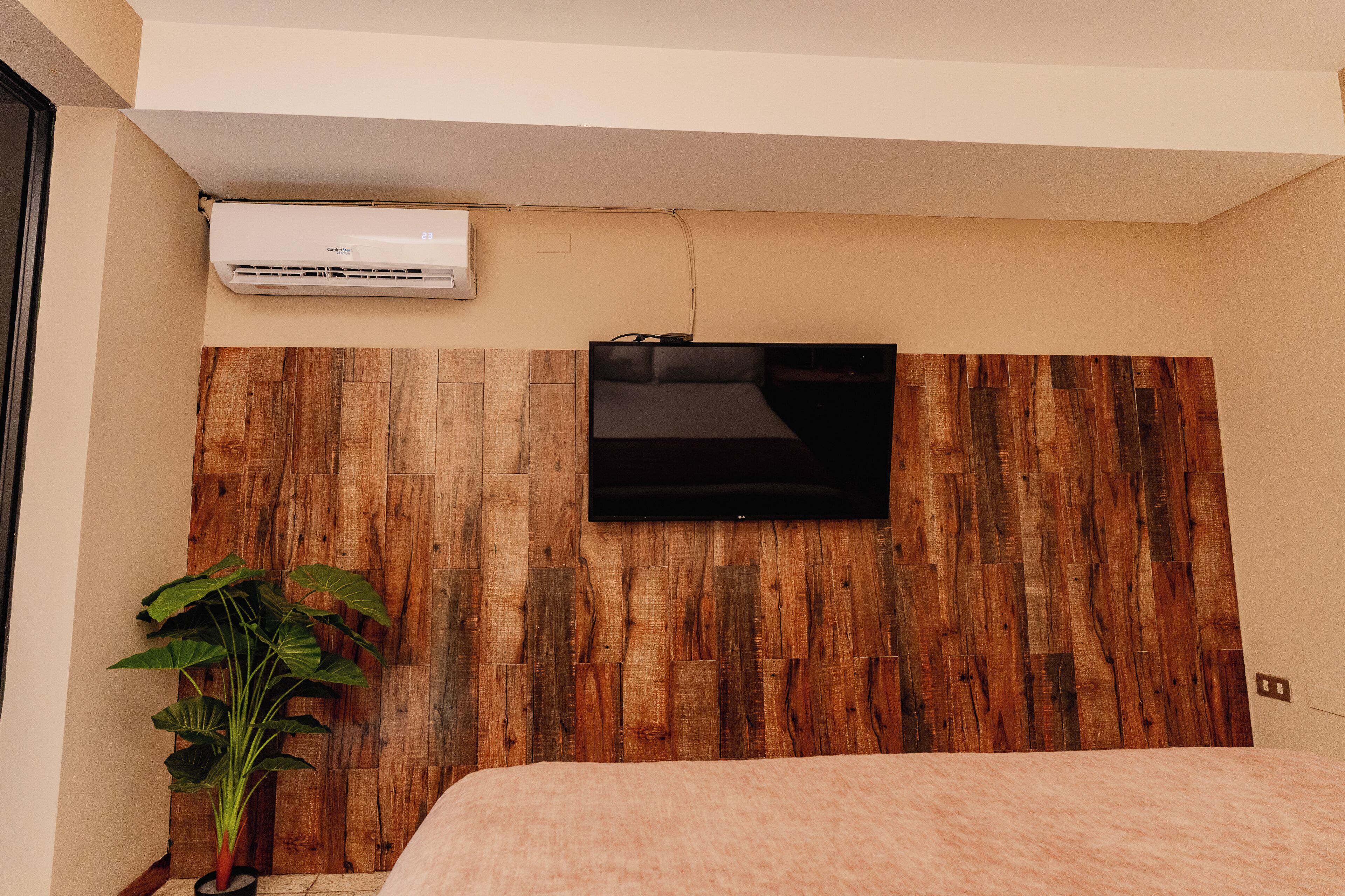 Ein Schlafzimmer mit Holz-Kopfteil, einem an der Wand befestigten Flachbildfernseher, einer Topfpflanze und einem Bett mit einer rosa gefärbten Decke.