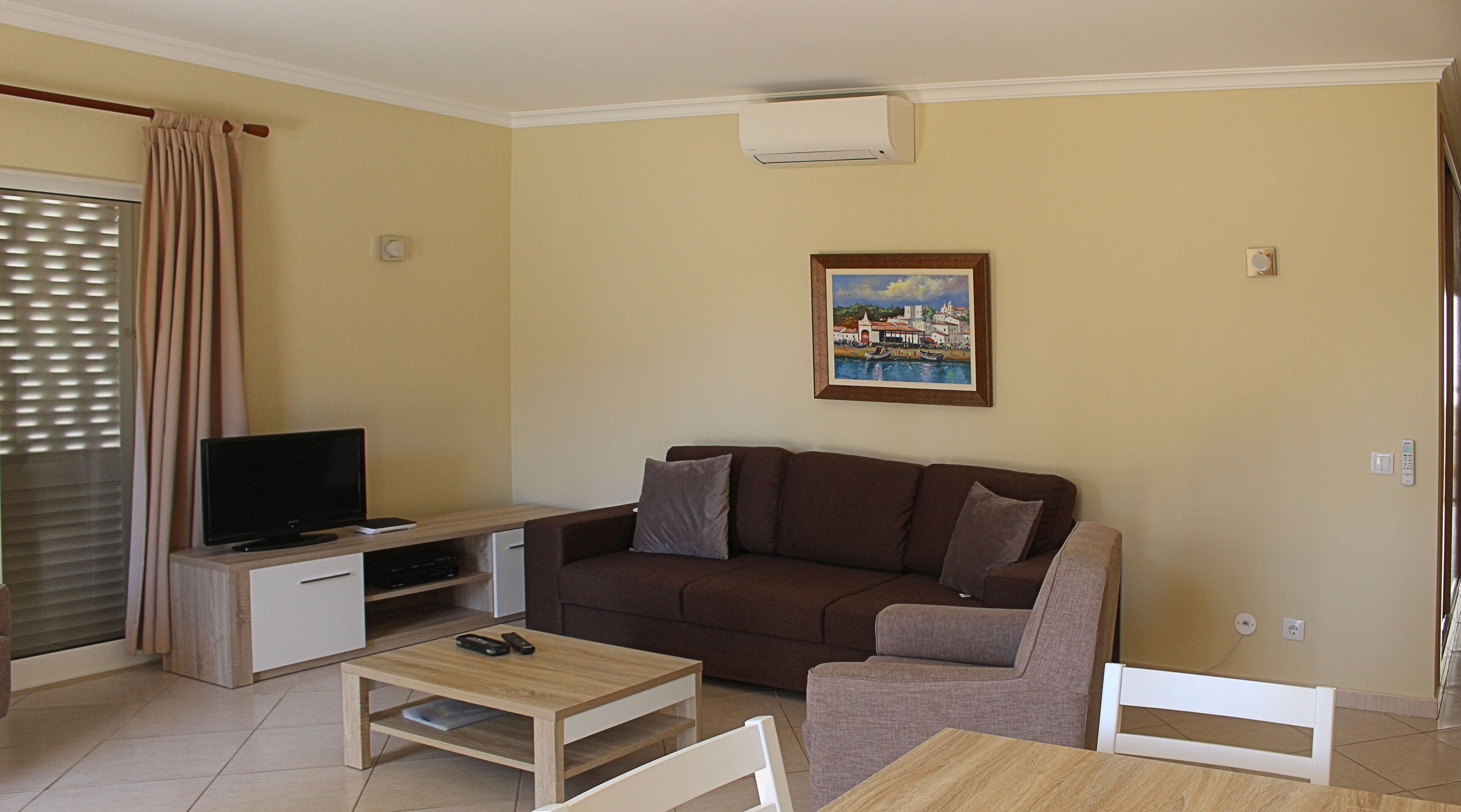 Una sala de estar con un sofá marrón, un televisor sobre un mueble de madera, una mesita y un cuadro en la pared.