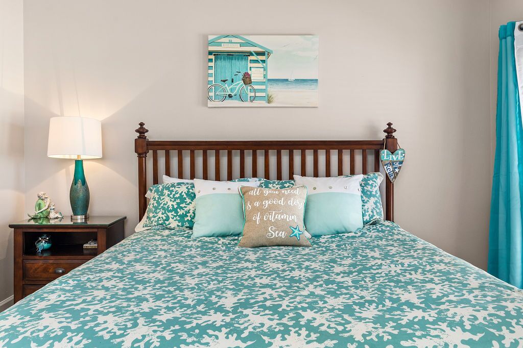 Een bed met een gebedekte spreideken, een nachtkastje met een lamp, een decoratief kussen, en een aan de muur hangend schilderij van een strandgezicht.