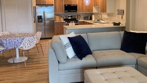 Una cocina moderna con armarios de madera, un refrigerador de acero inoxidable y un horno empotrado. En la sala de estar, hay una mesa de comedor con sillas blancas y un sofá de color claro.