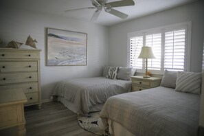 Une chambre avec deux lits, une commode, un ventilateur de plafond, un tableau et une fenêtre avec des stores.