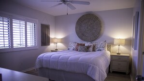 A bedroom with a large bed, two bedside tables with lamps, a ceiling fan, a large wall decoration, and a window with blinds.