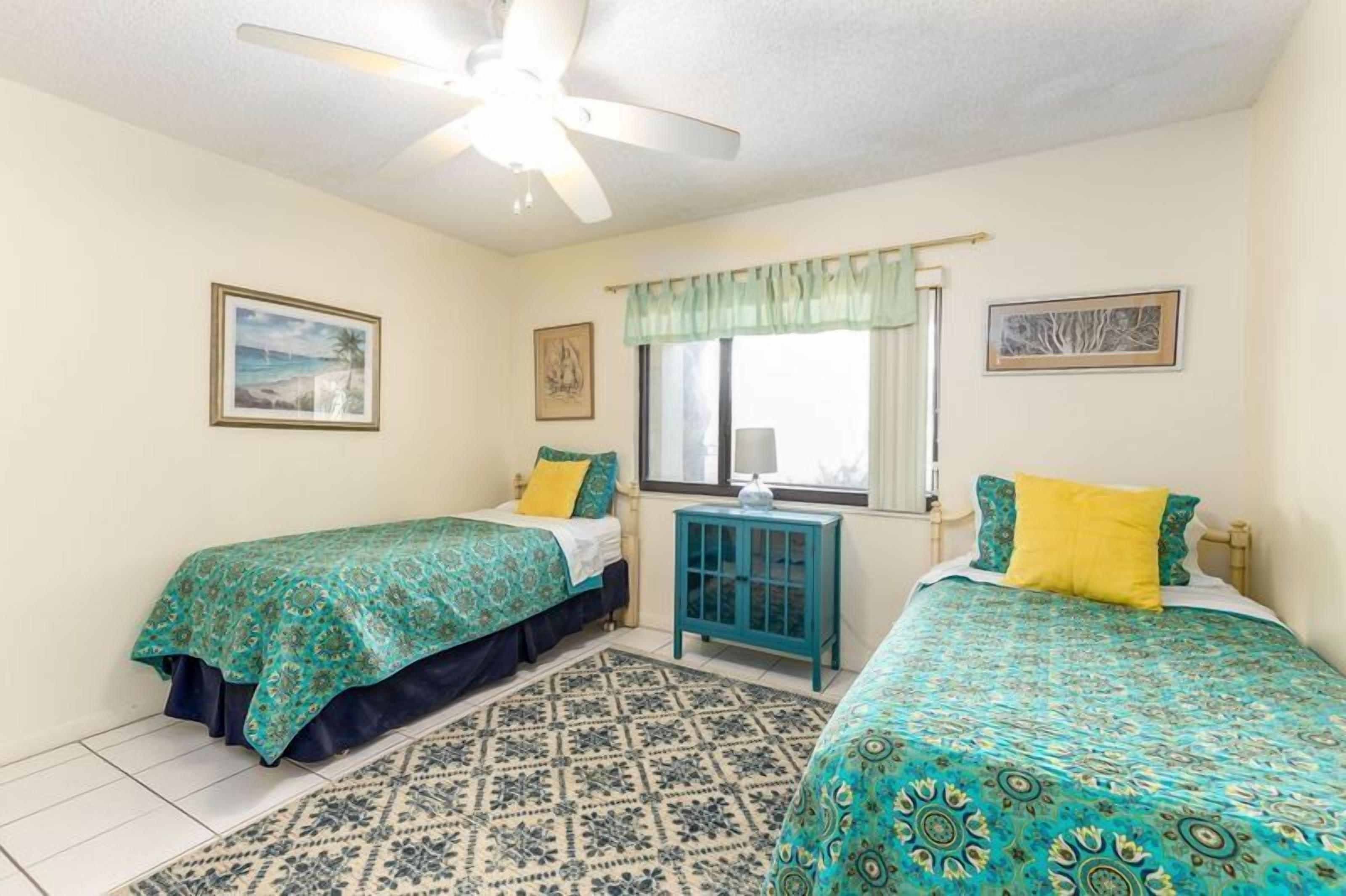 Une chambre avec deux lits, un ventilateur de plafond, une fenêtre avec un rideau et un tapis à motifs.
