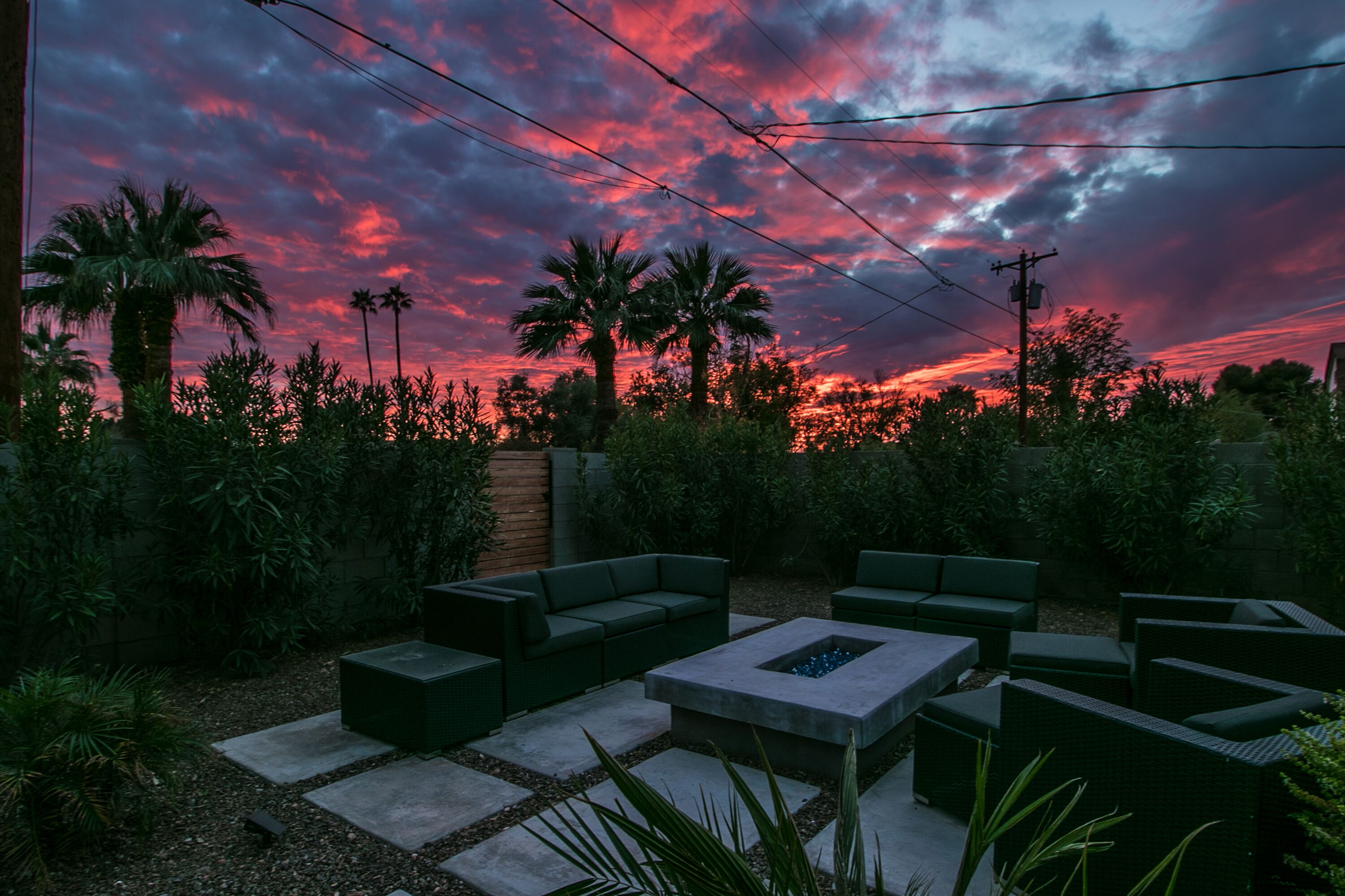 Une terrasse dotée de mobilier d’extérieur et d’un foyer, entourée de verdure et de palmiers, sous un ciel spectaculaire.