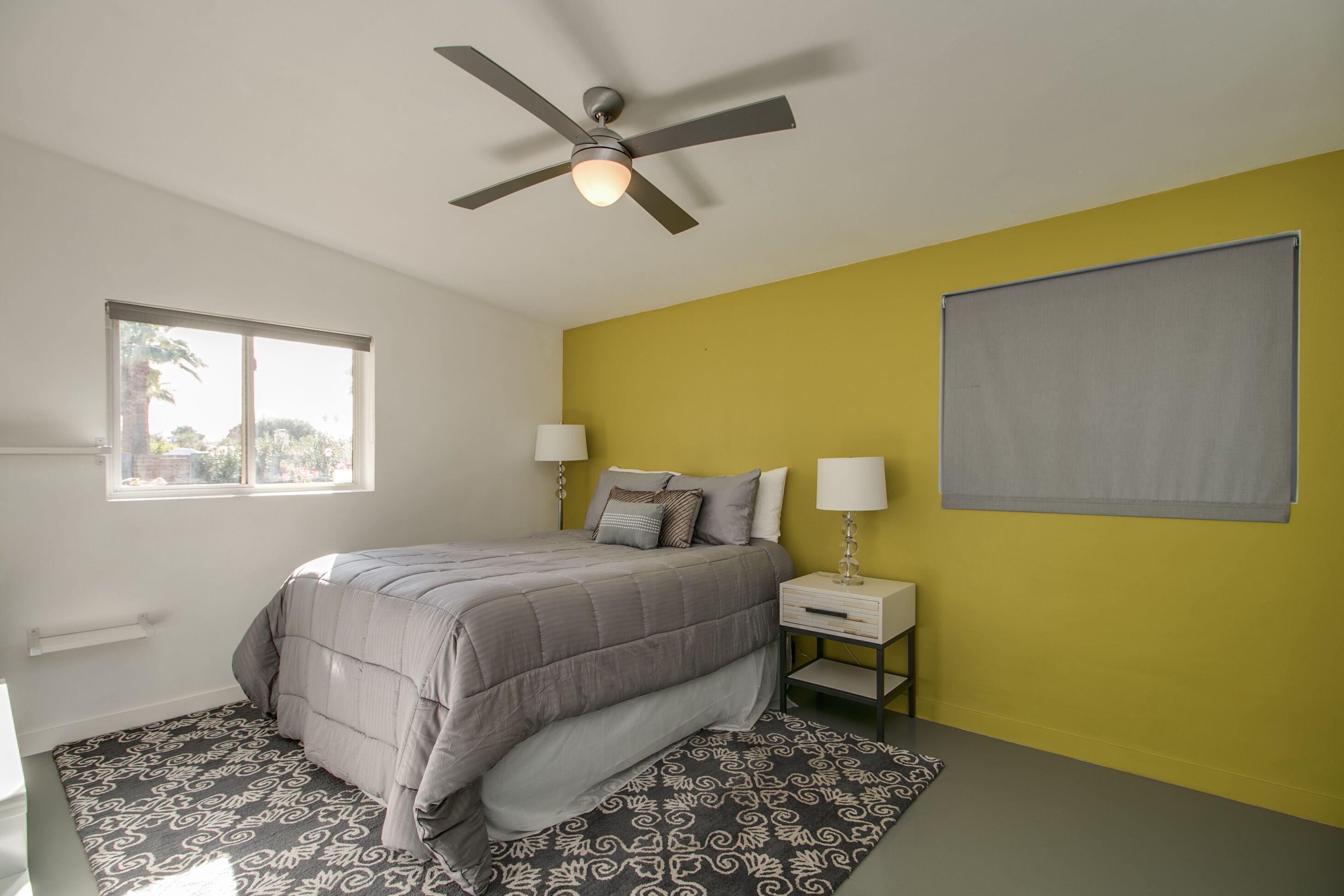 A bedroom with a bed, bedside tables, a ceiling fan, a window, and a wall-mounted light fixture.
