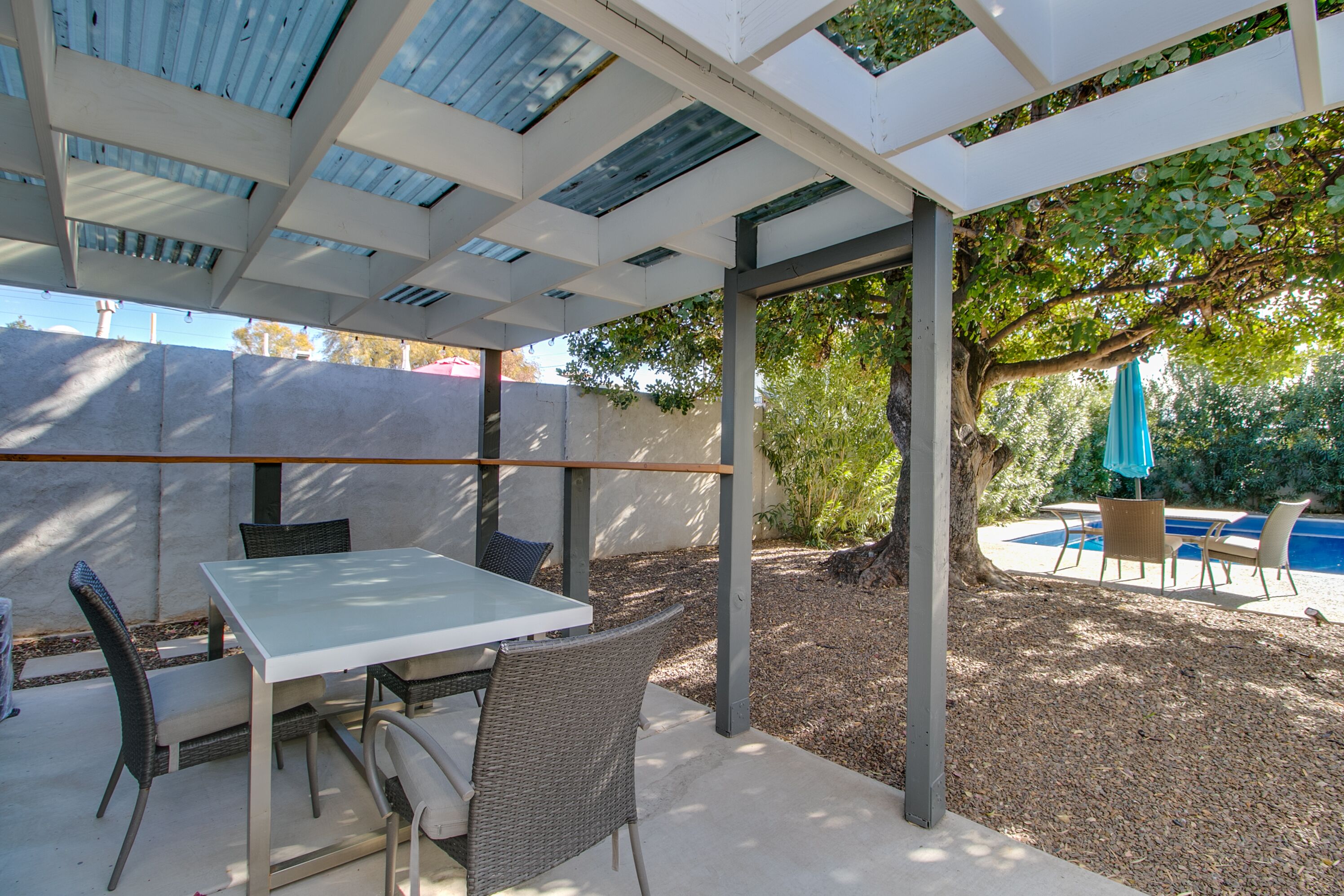 An outdoor patio with a table and chairs, a pergola with a roof, and a view of a swimming pool.