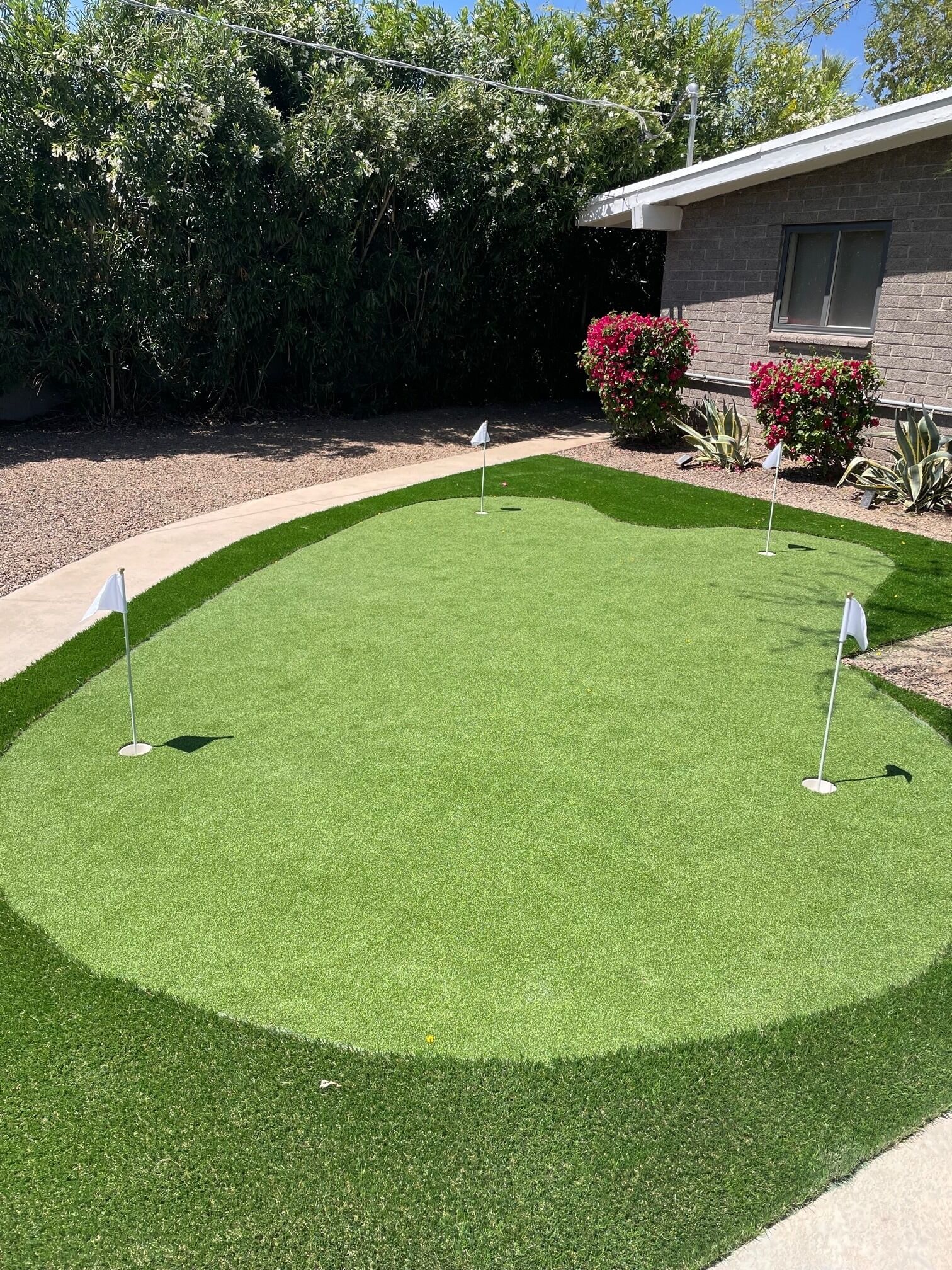 Un área de práctica de golf artificial bien cuidada, con tres banderas, rodeada de un jardín paisajístico y un edificio al fondo.