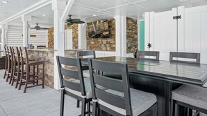 Un espace extérieur aménagé pour les repas, avec un mur en pierre, un téléviseur fixé au mur et un ventilateur de plafond.