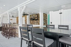 An outdoor dining area with a stone wall, a TV mounted on the wall, and a ceiling fan.