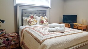 A neatly made bed with a floral-patterned bedspread and two folded towels, a television on a wooden stand, and a suitcase with a South Africa map design. - Lower Suite Duplex - 3 Beds / 1 Bath By  Restaurants, Costco, G P Airport  (Grande Prairie)