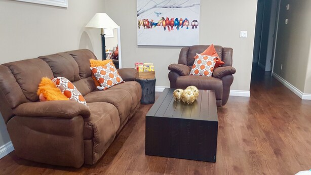 A living room with a brown sofa, a brown armchair, and a coffee table. There is a lamp, a picture on the wall, and a small side table with a box of tissues. - Lower Suite Duplex - 3 Beds / 1 Bath By  Restaurants, Costco, G P Airport  (Grande Prairie)