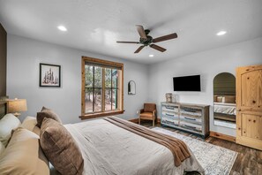 Une chambre à coucher comprenant un lit, une chaise, une commode, un ventilateur de plafond, un téléviseur et une grande fenêtre.