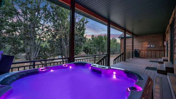 A hot tub with purple lighting on a wooden deck with a view of trees and a building in the background. - East Zion Trails Retreat: Your Gateway to Zion (Orderville)