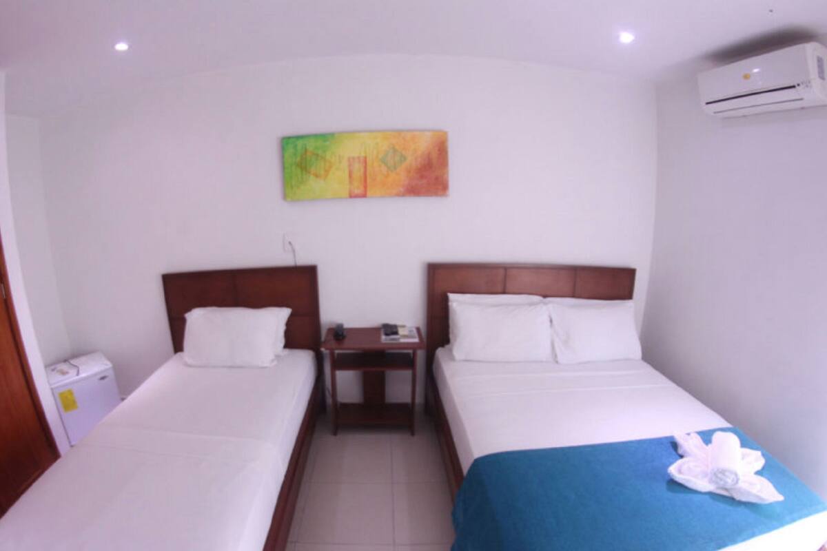 A hotel room with two single beds, a wooden wardrobe, a small table with a chair, an air conditioning unit, and a colorful painting on the wall.