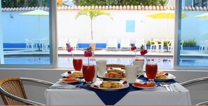 Une table de petit-déjeuner avec des assiettes remplies de nourriture, des boissons, et une vue sur un espace piscine avec des chaises et des parasols.
