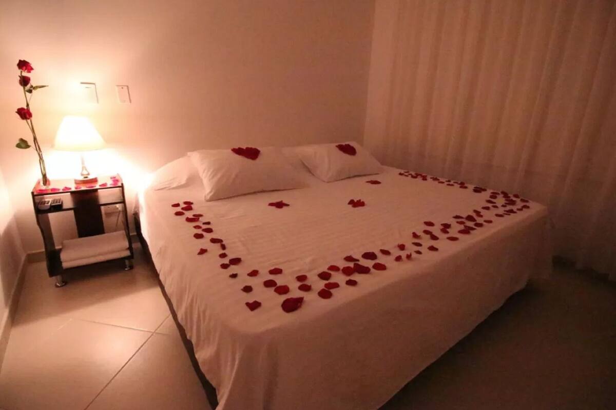 A neatly made bed with rose petals and heart-shaped pillows, a bedside table with a lamp, and a curtain in the background.