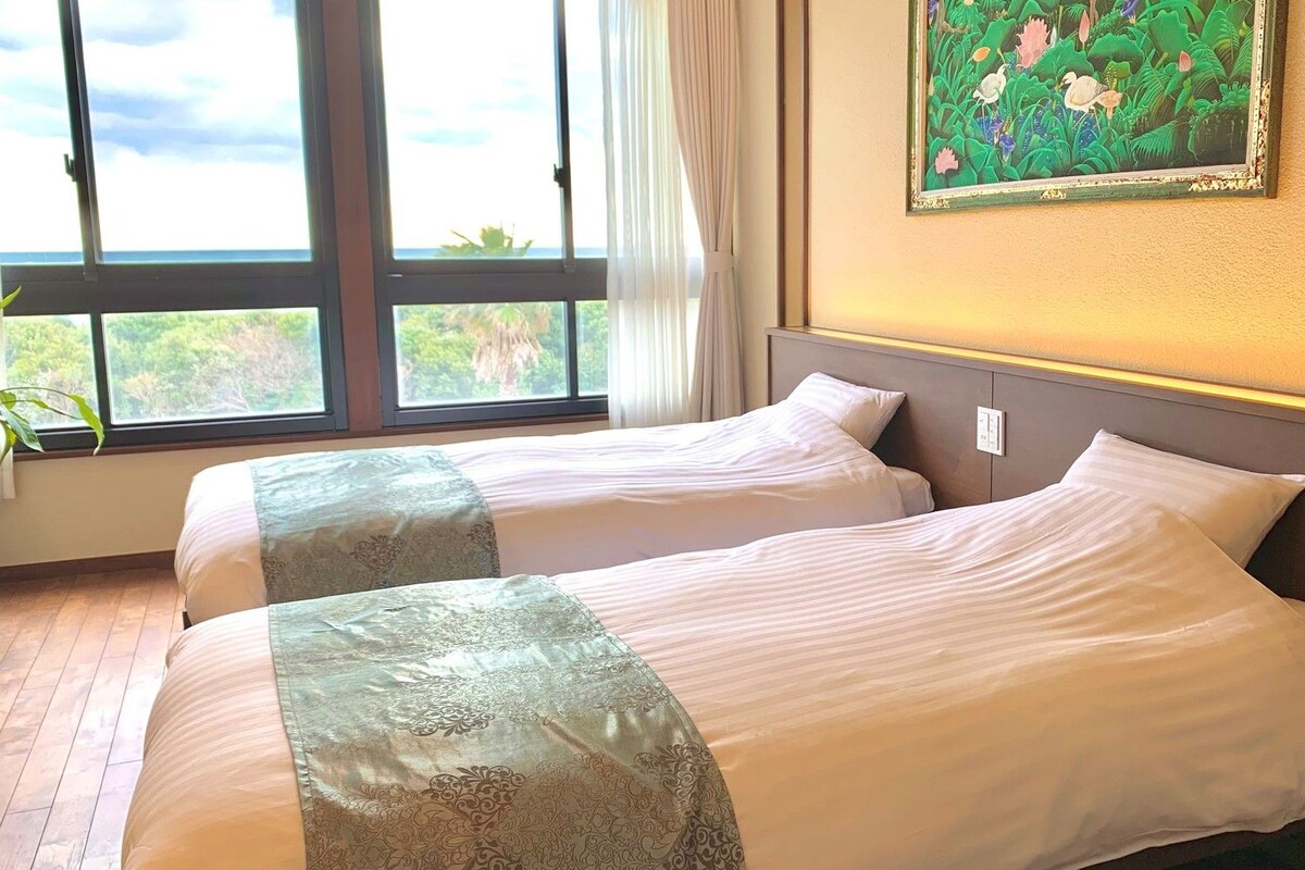 A hotel room with two beds, a large window with a view of greenery, and a painting on the wall.