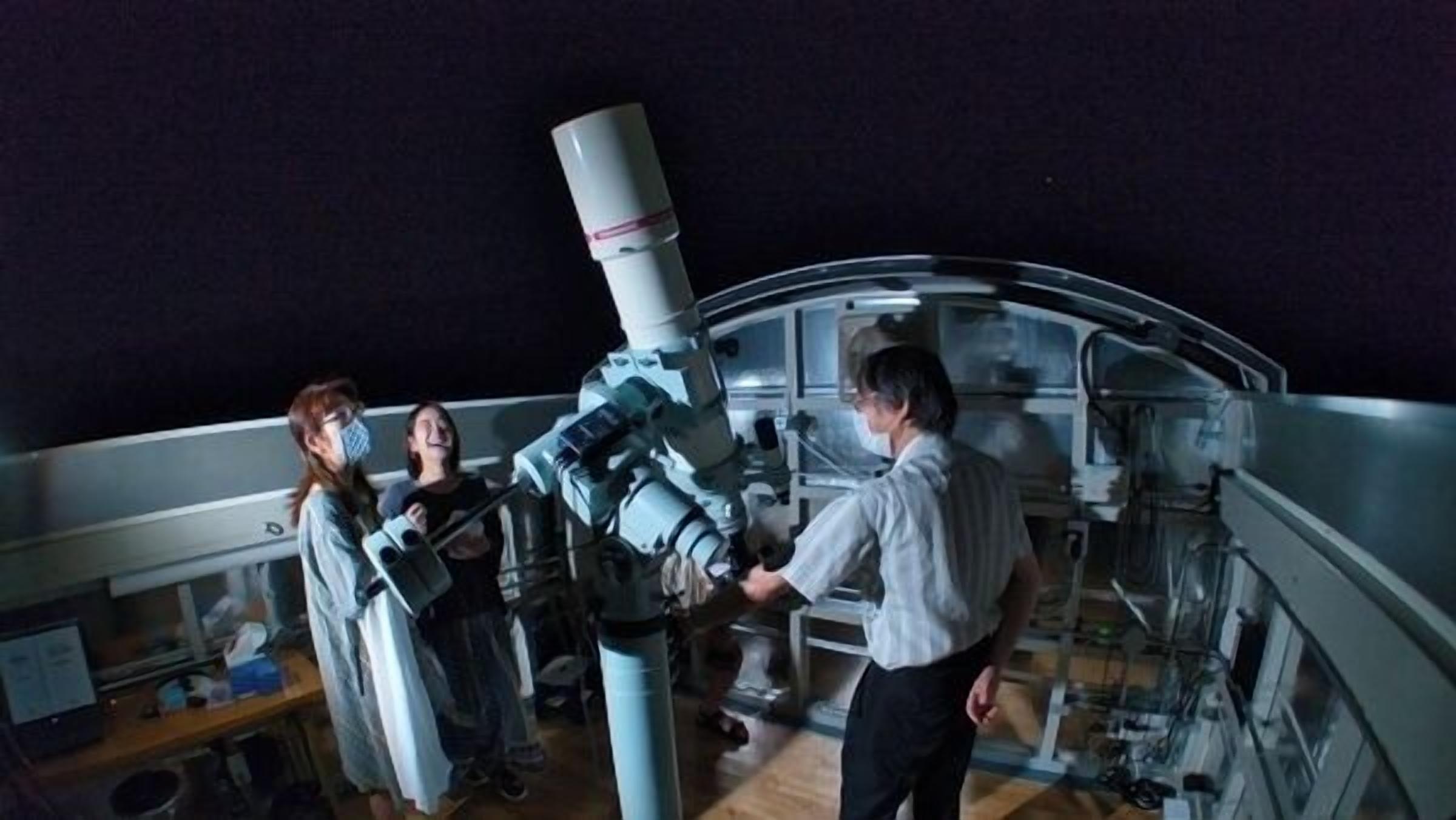 A telescope in a room with a high ceiling and a starry sky visible through the windows.