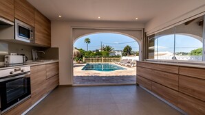 A modern kitchen with wooden cabinets, a built-in microwave, and an oven. It leads to a pool area with a view of the surrounding landscape. - Villa for 7 (Xabia)