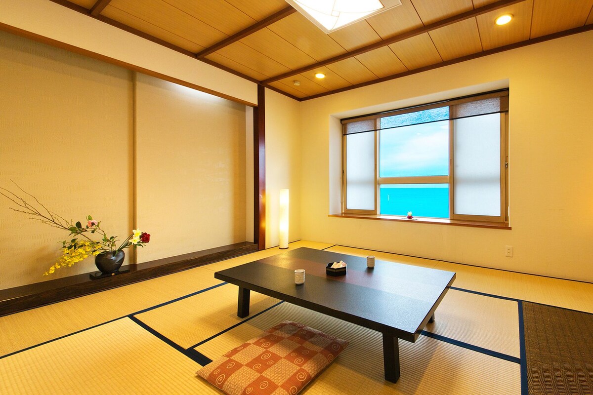 A traditional Japanese room with tatami flooring, a low table, a window with a view, and a vase with flowers.