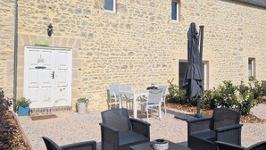 Un edificio de piedra con una puerta blanca, mobiliario exterior con sillas y mesas, y una sombrilla cubierta.