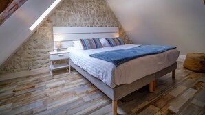 Un dormitorio con techo inclinado, pared de piedra, suelo de madera, cama blanca, almohadones a rayas y una lámpara colgante.