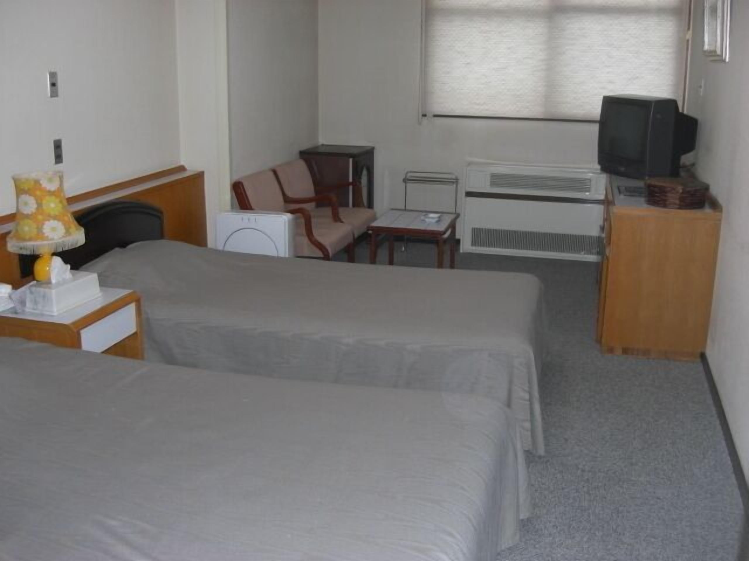 A hotel room with two beds, a desk with a lamp, a chair, a television, and a radiator.