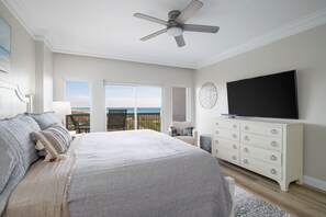 Une chambre dotée d’un grand lit, d’un ventilateur de plafond, d’une télévision et offrant une vue sur l’océan par les fenêtres.