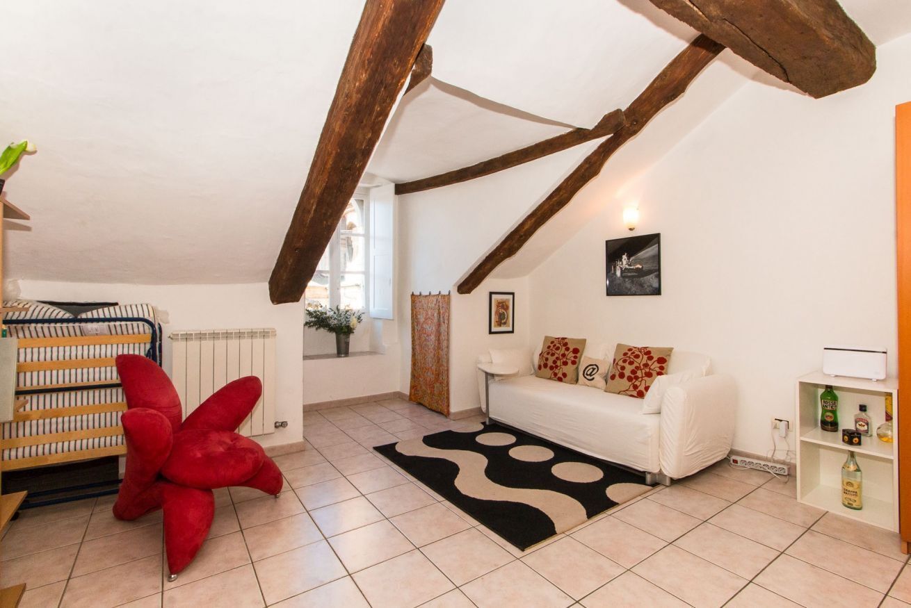 A cozy loft-style room with exposed wooden beams, a white sofa set, a large red bean bag, and a black and white patterned rug.