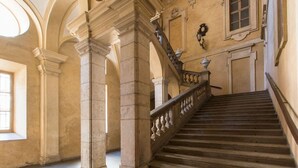 Un magnifique escalier en pierre, doté de balustrades, mène à l’étage supérieur d’un bâtiment à l’architecture classique, caractérisé par des arches et des colonnes.
